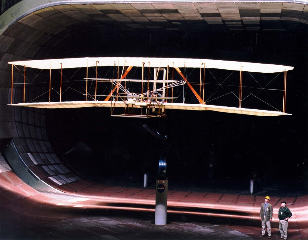 A full-size Wright Flyer replica mounted on a test stand inside the Ames wind tunnel, with two engineers standing beside it.